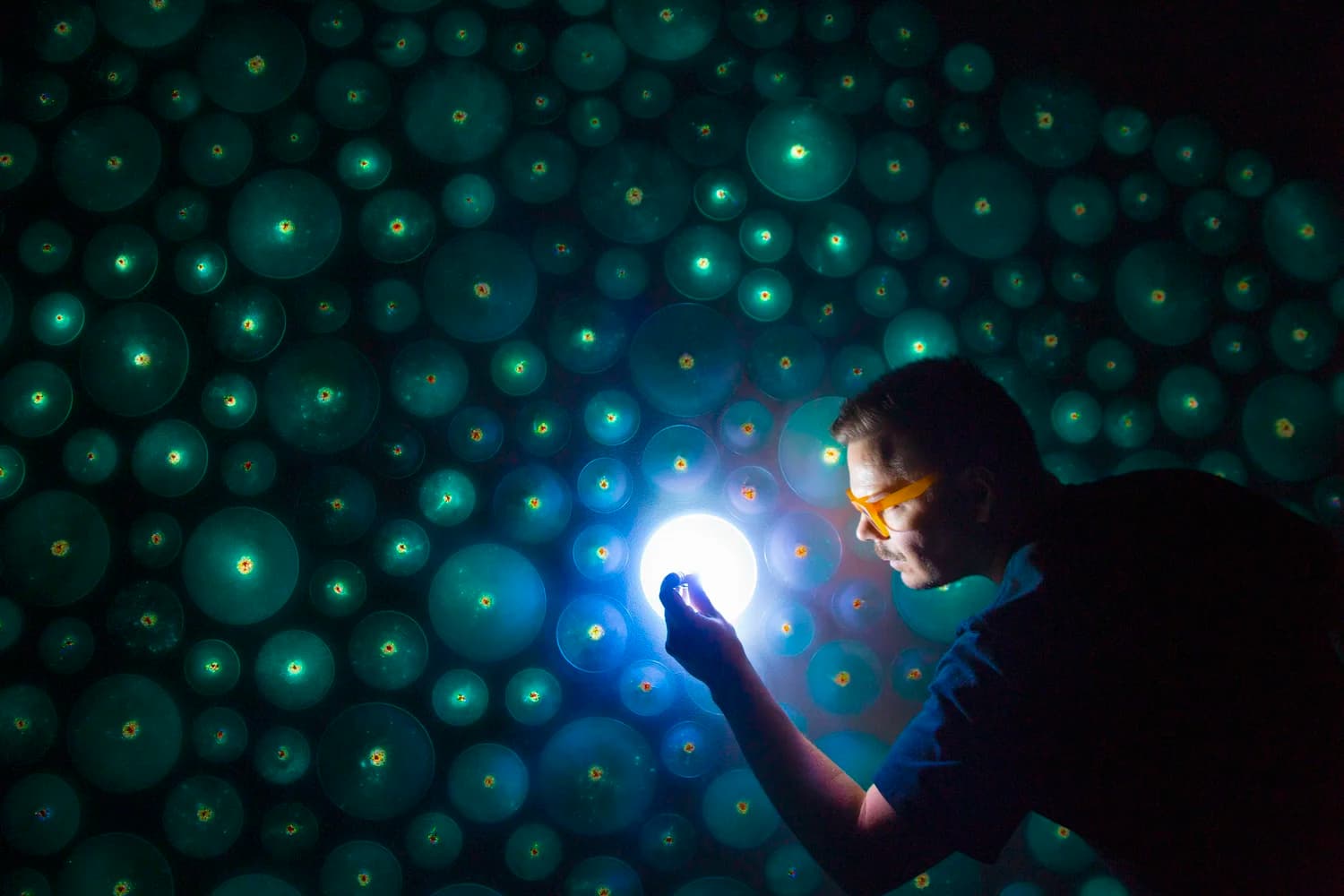 Close-up — the artist's hand cradles a glowing orb against a wall of softly lit teal spheres, each one pinpricked with a brighter core.