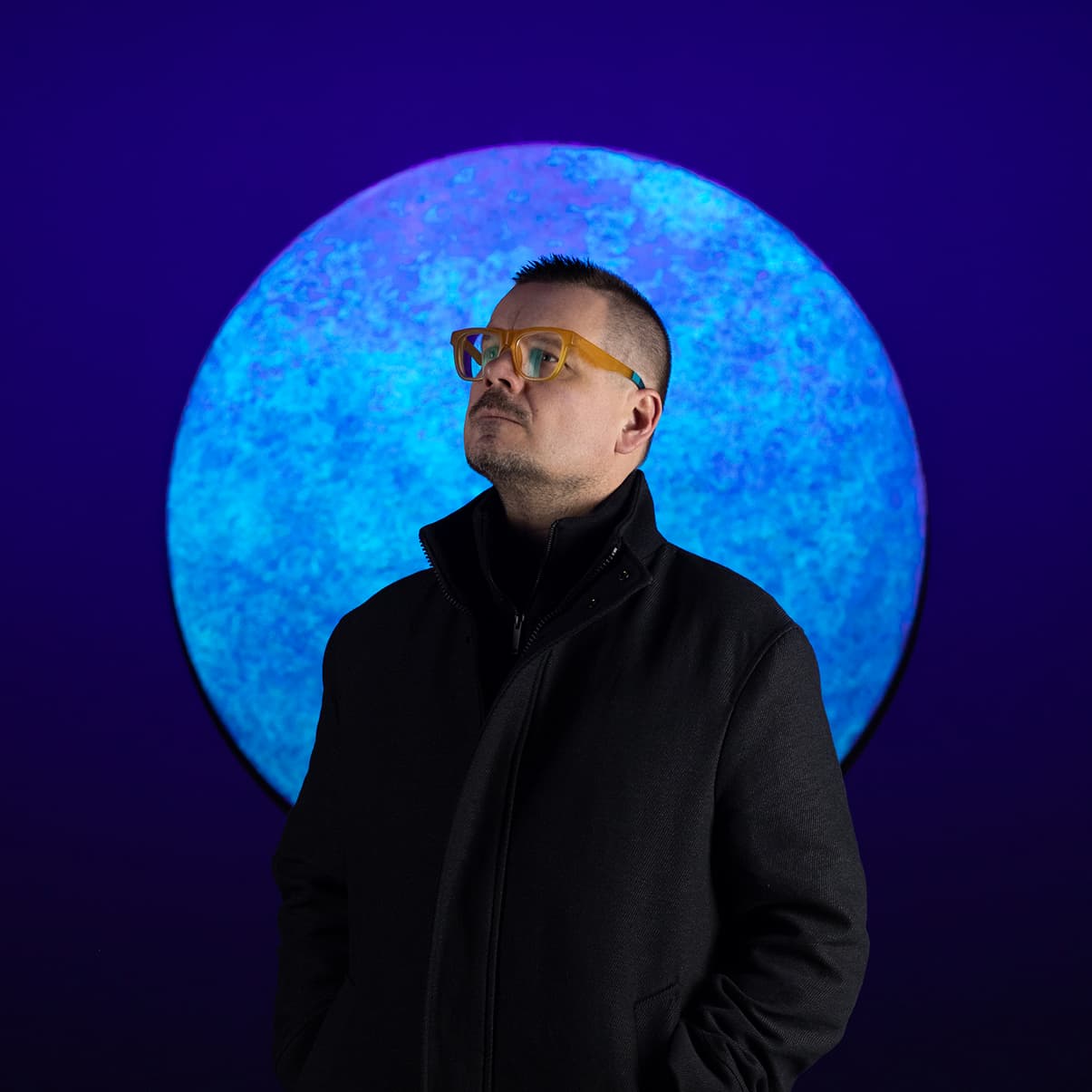 Portrait of Cristopher Cichocki standing before a large glowing blue moon-like sphere.