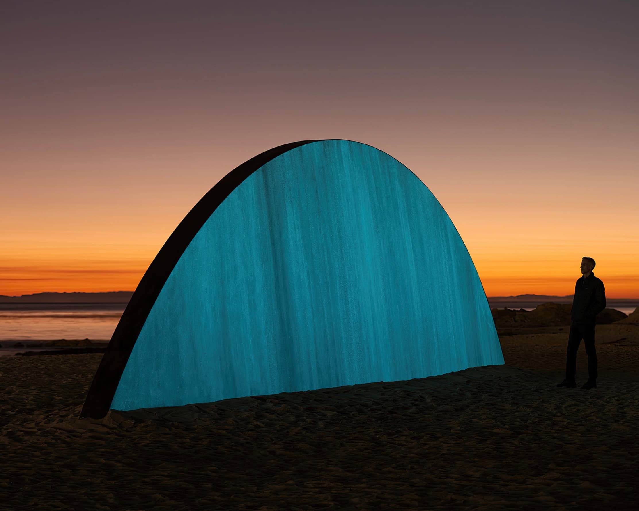 A large luminous teal half-dome sculpture on a darkening beach at sunset, a lone figure silhouetted beside it against an orange horizon.