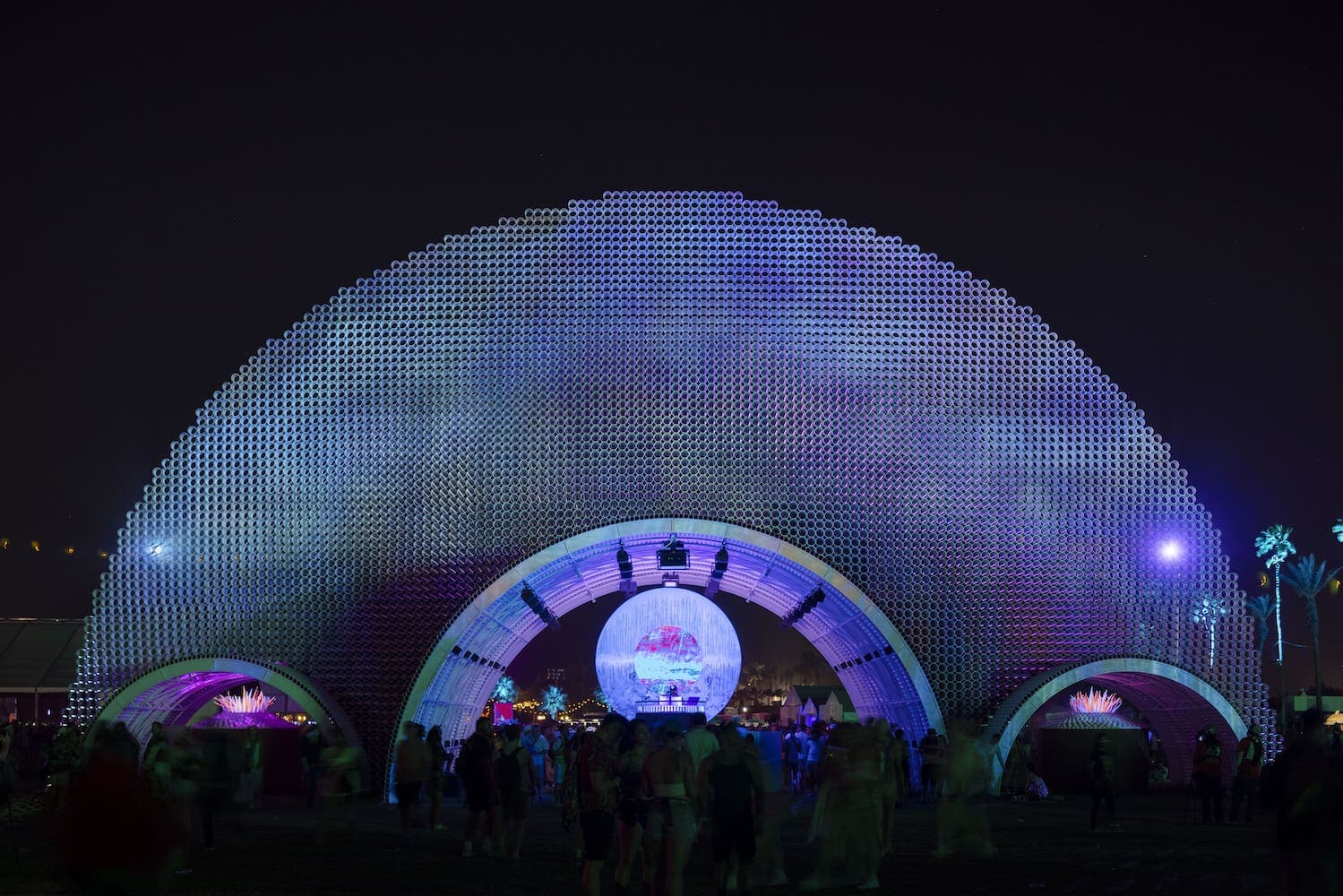 A massive luminous dome made from thousands of PVC tubes glowing white and lavender against the night sky, surrounded by a crowd at the Coachella festival.