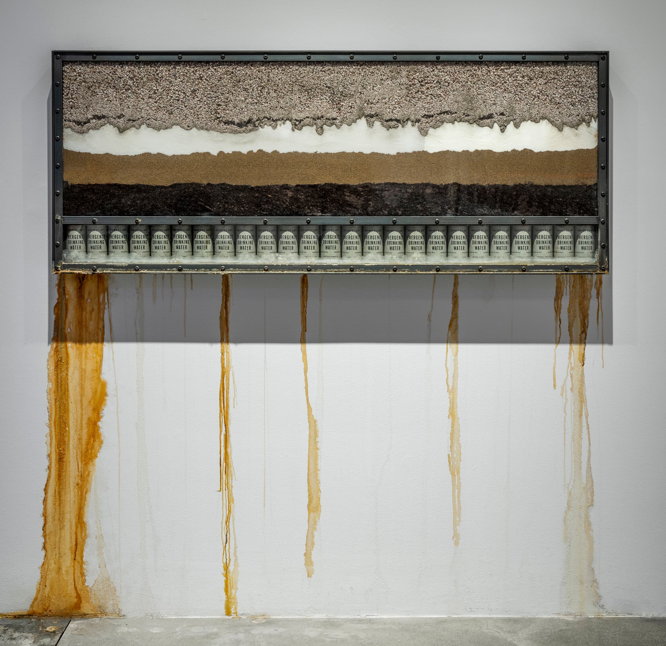 A wall-mounted vitrine of layered mineral sediments — black, white, and ochre — leaks long mineral-coloured streams of water down the gallery wall and pools on the floor.