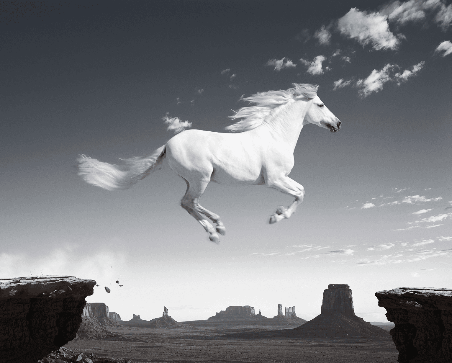 Black-and-white photograph of a white horse mid-leap across a desert chasm, with Monument Valley buttes on the horizon.